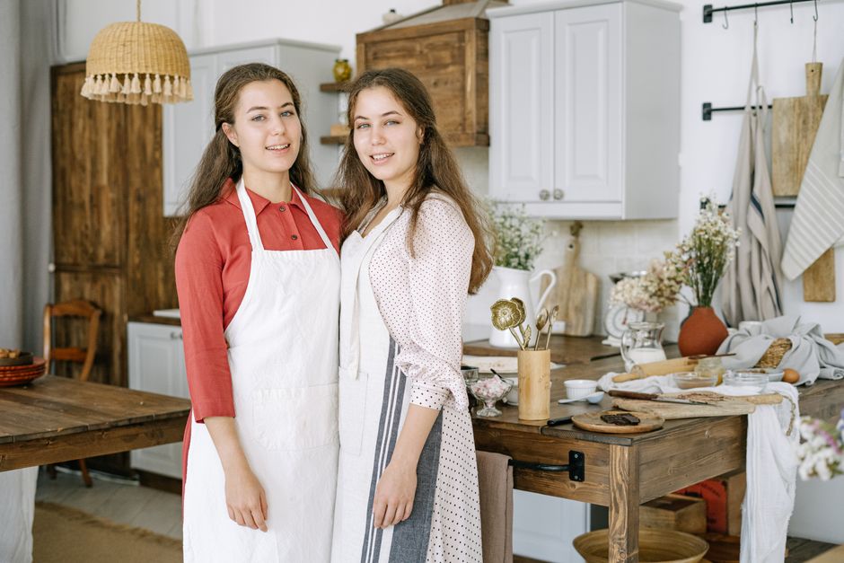 Two young women wearing aprons in a cozy kitchen, smiling and posing together.