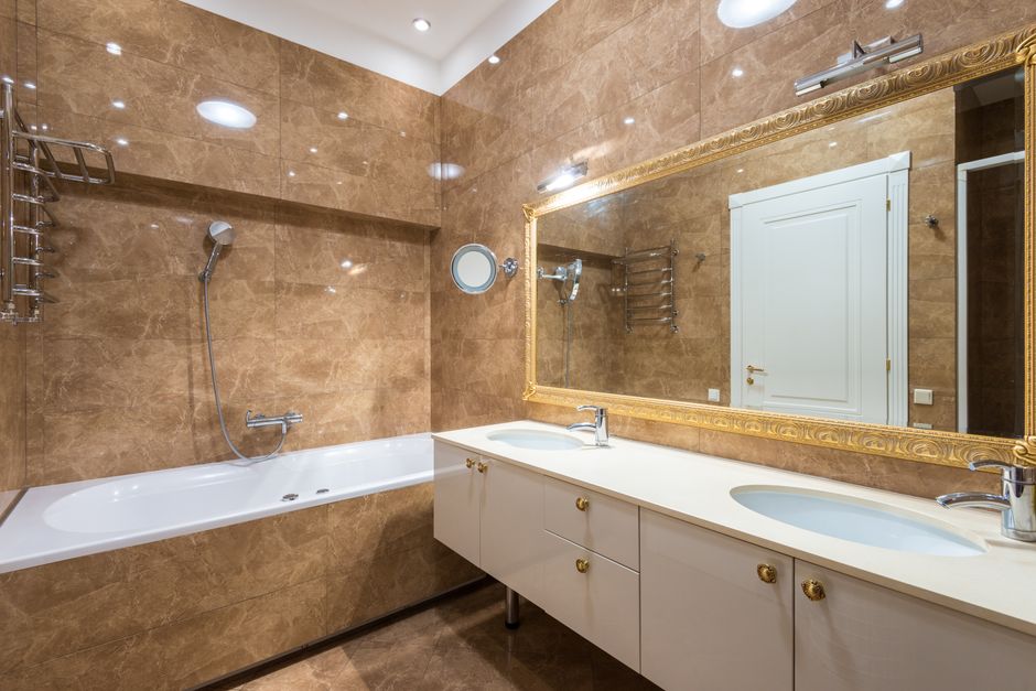 Interior of bathroom with wide mirror hanging above sinks near bathtub and shower on marble tiled wall