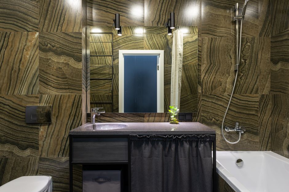 Stylish bathroom with tiled walls furnished with bath and sink with toilet under glowing lamps