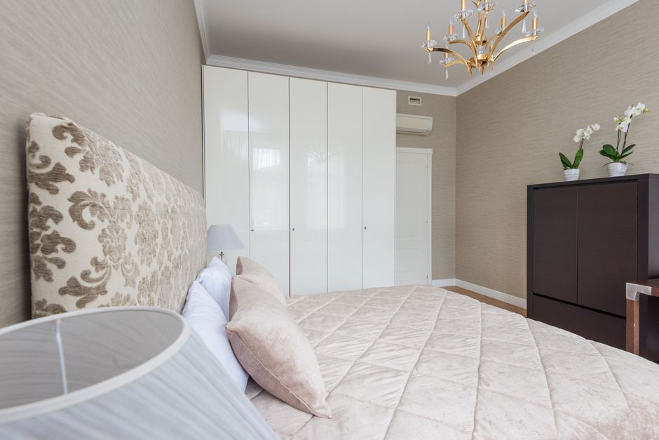 Bed under coverlet with cushions placed near lamp on bedside and closet near door in bedroom with classic chandelier