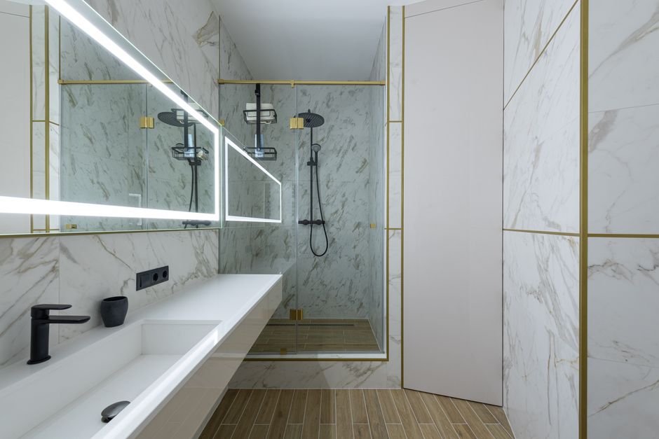 Shower cabin and ceramic sink in spacious modern bathroom with marble tiled walls and big window with bright lamps
