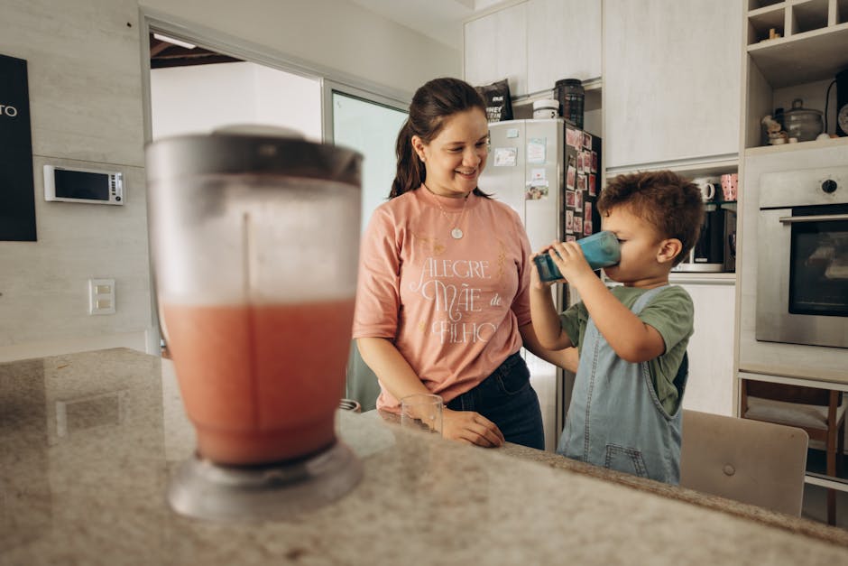 A mother smiles as her son drinks a smoothie from a blue cup in the kitchen, conveying family bonding.