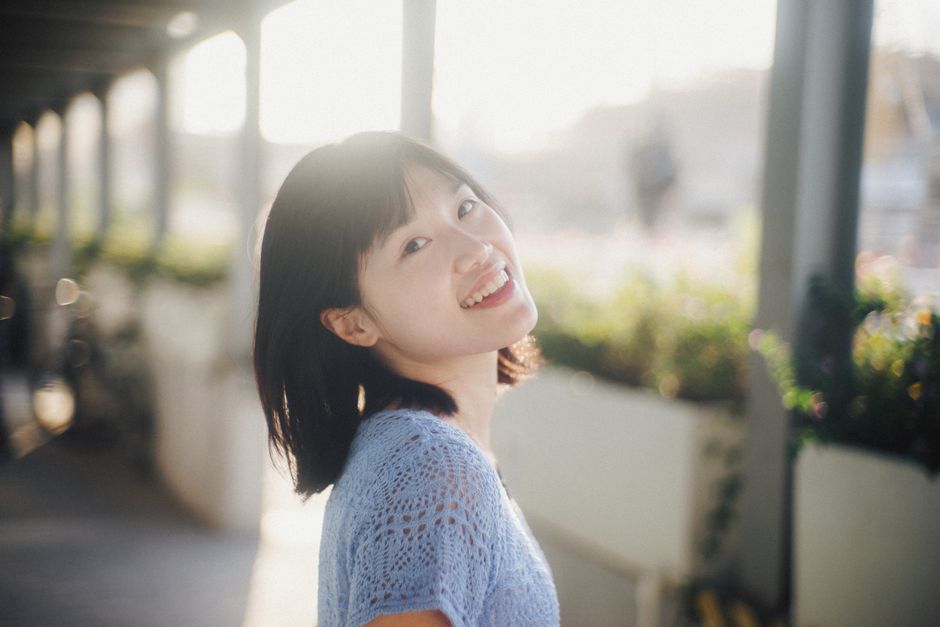 A bright and joyful portrait of a smiling young woman standing outdoors in sunlight.