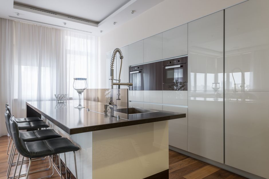 Leather chairs placed at island in modern spacious kitchen with minimalist white shiny furniture and built in appliances in daylight