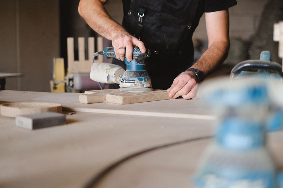 Unrecognizable male woodworker wearing uniform and polishing wooden board with random orbital sander at big table in professional studio