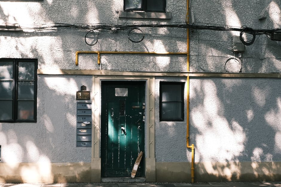 Exterior wall with green door, shadows, and urban elements including pipes and wires.