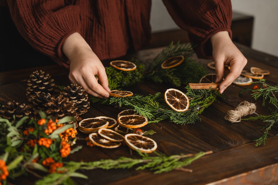 Hands crafting a Christmas wreath with dried oranges, pinecones, and greenery.