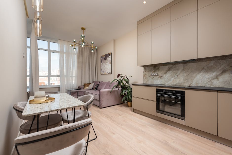 Interior of modern light studio with open kitchen with cabinets near table and chairs next to couch with pillows under chandelier near windows with curtains