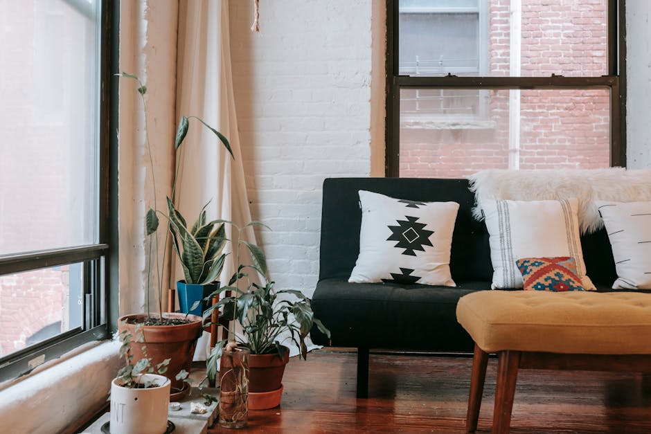 Part of comfortable sofa with yellow soft bench placed near windows and potted green plants on floor in modern apartment