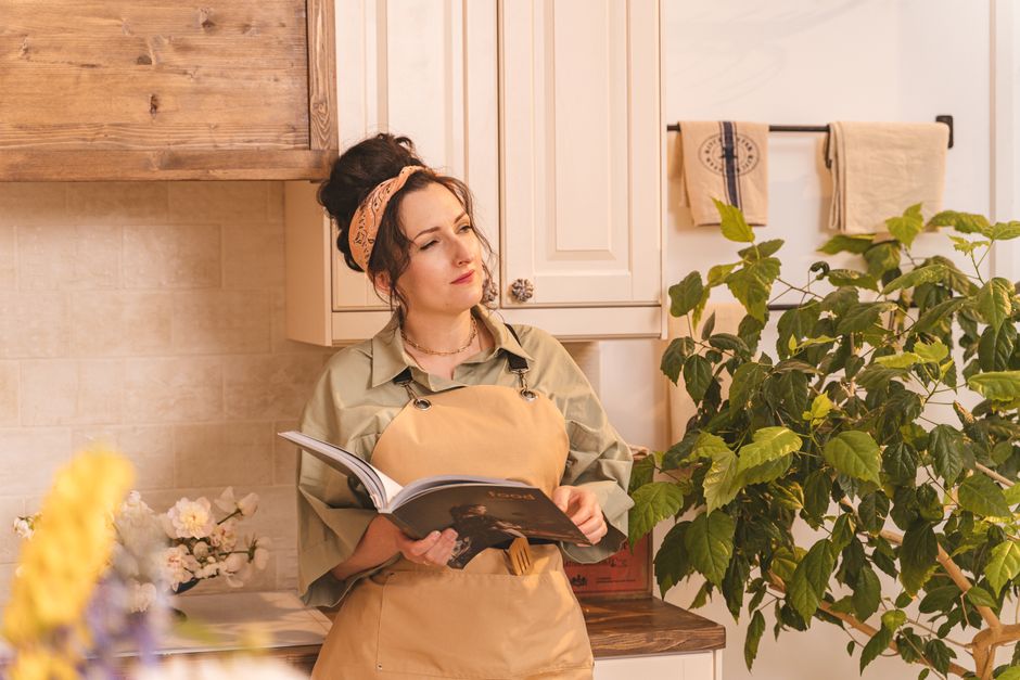 Woman with bandana and apron reads a book in a warmly lit, plant-filled kitchen.