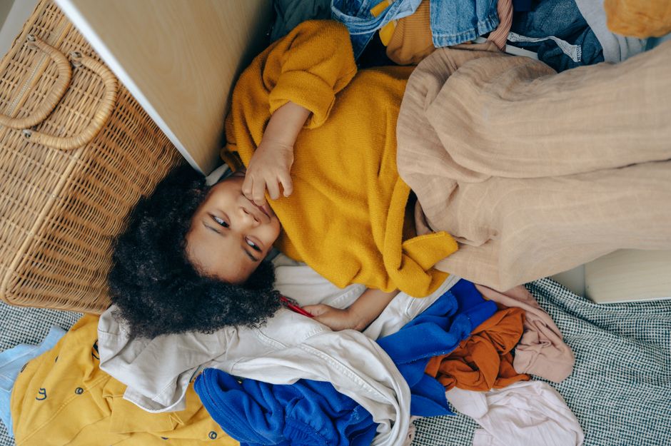 From above of playful little black girl smiling and lying on pile of various clothes after making mess in room