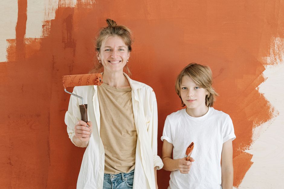 A mother and son smiling while painting a wall indoors, holding paint roller and brush.
