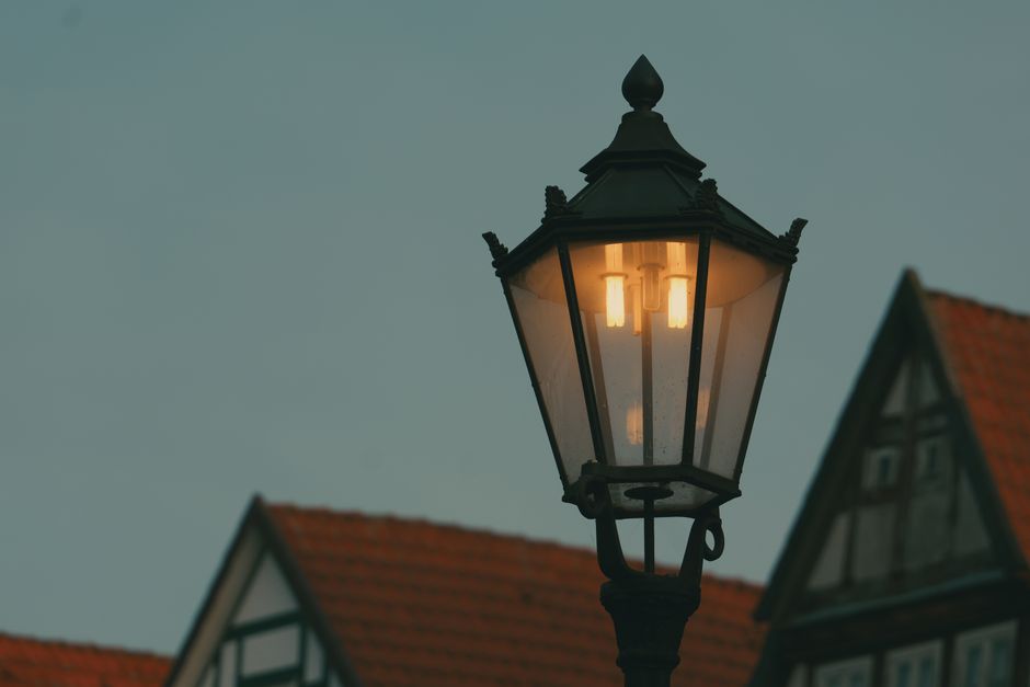 An atmospheric image of a vintage street lamp glowing against traditional houses with red roofs at twilight.