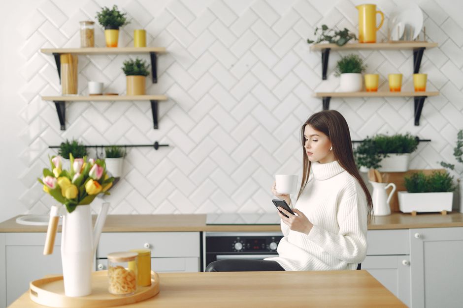 Side view of focused young woman in warm sweater browsing mobile phone in cozy kitchen and drinking hot beverage while resting at table with bouquet of tulips in kitchen