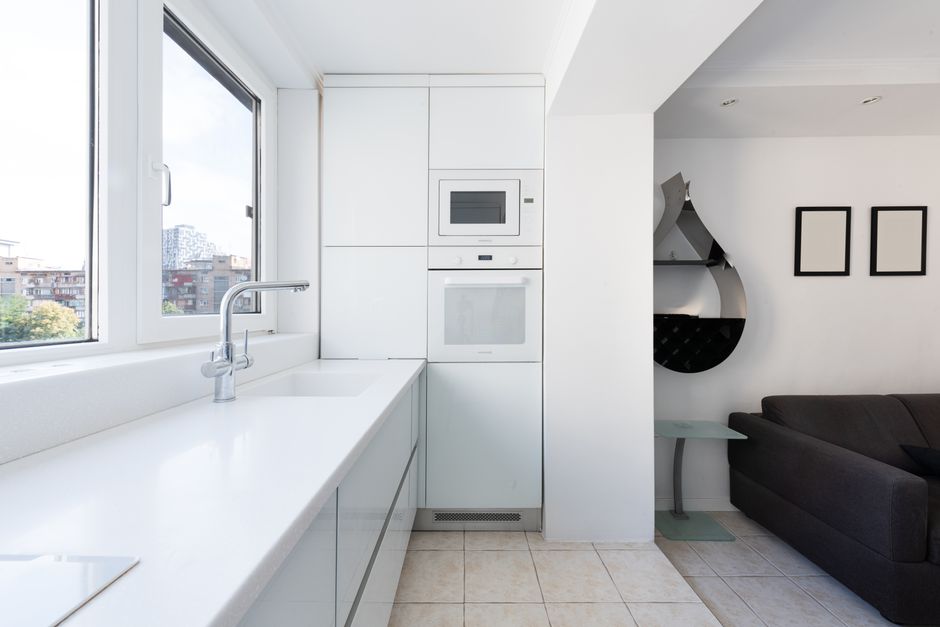 Modern studio interior with kitchen cabinets with sink and tap near oven and microwave near windows and living room with sofa near table and shelves on wall in daylight