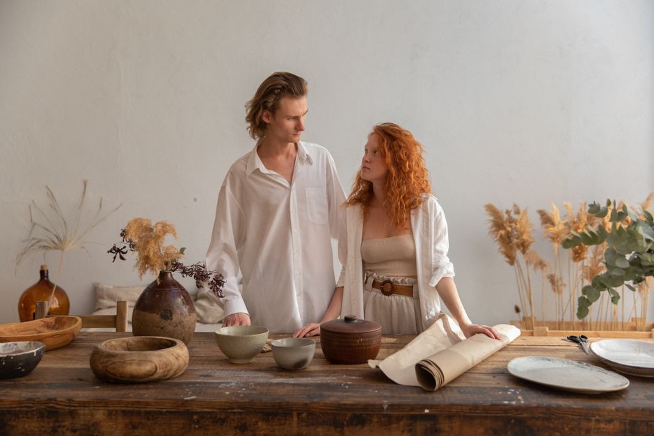 Young charismatic couple in stylish clothes standing near wooden table with various utensils for crafting and looking at each other